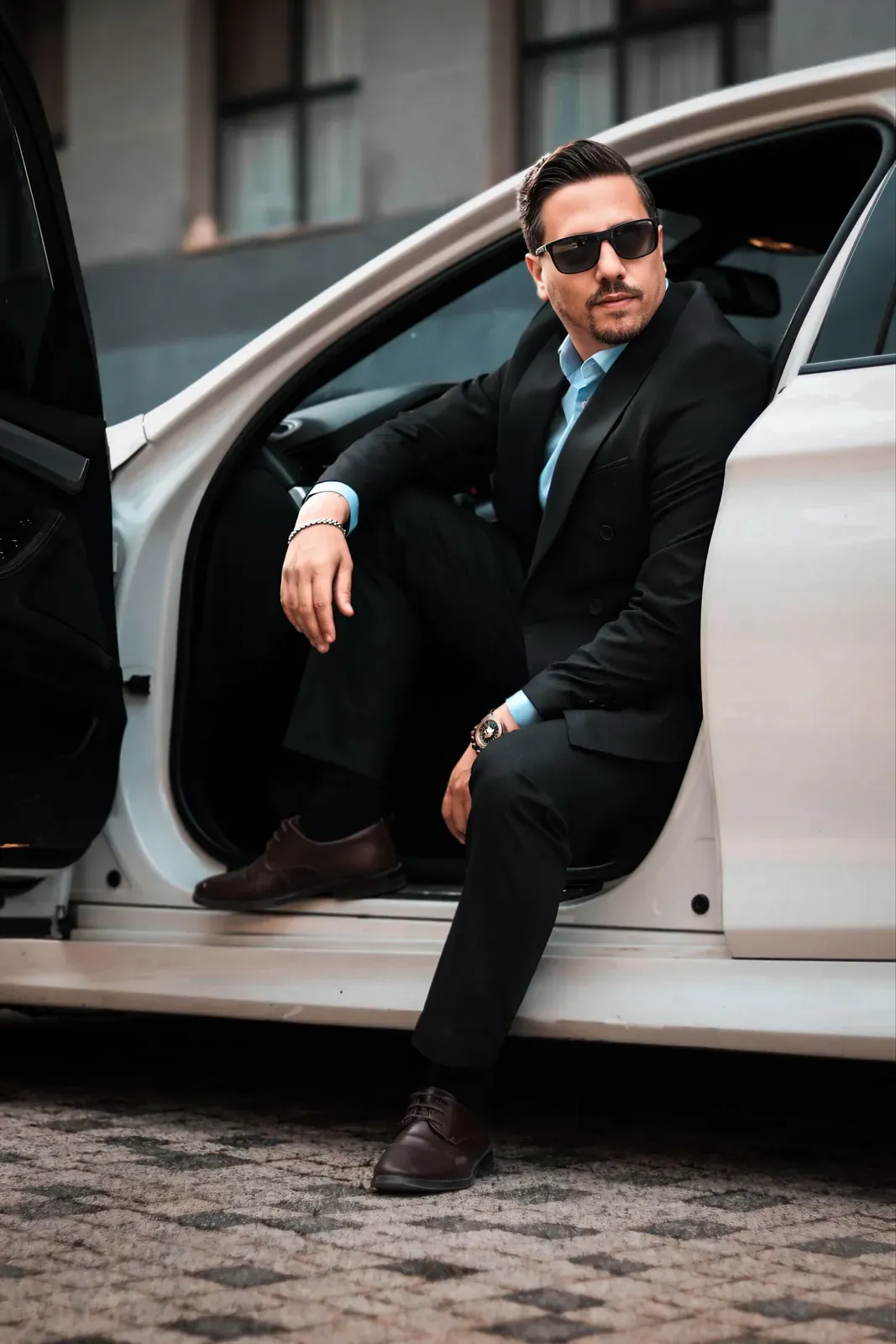 A man in a dark suit seated in the open door of a car