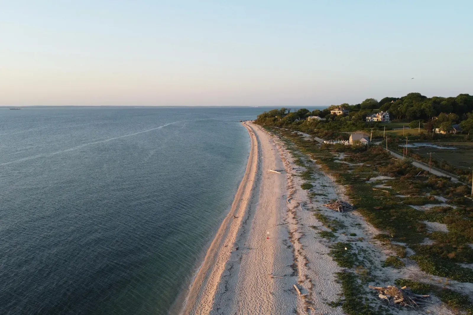 Aerial view of a long, narrow beach beside calm water at sunset, with houses on the bluff